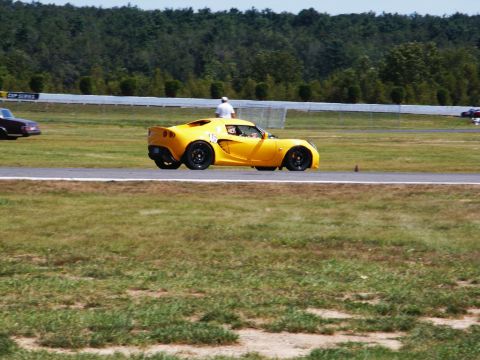 2001 Mercury Grand Marquis - A yellow Lotus Elise with racing number 16 navigates a turn on a sunny day at the track, with another car visible in the background.