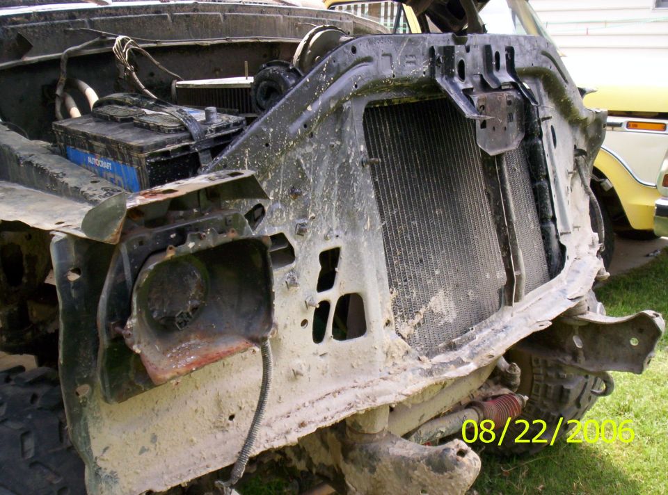 Front end of a heavily damaged truck, showing the radiator and battery. Another yellow truck is visible in the background