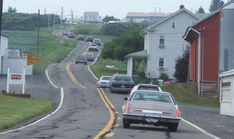 1985 Ford LTD Crown Victoria - A cruise convoy of full-size sedans strung out along a hilly two-lane road through a small farm town, shot over the trunk of a silver Ford…