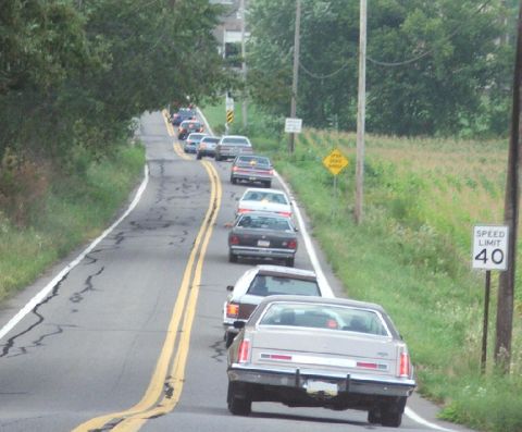 1985 Ford LTD Crown Victoria - A long line of full-size American sedans parked nose-to-tail along the shoulder of a rural two-lane road, shot from behind at a car-club cr…