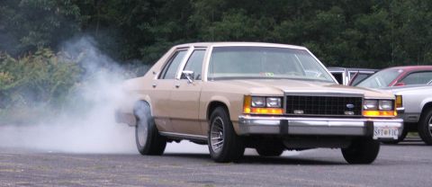 1985 Ford LTD Crown Victoria - A tan 1985 Ford Crown Victoria LTD is captured mid-burnout, emitting a large cloud of smoke. Another vehicle is visible in the background.