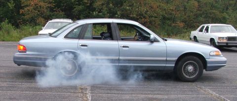 1985 Ford LTD Crown Victoria - Side profile of a light blue Ford Crown Victoria sedan doing a burnout in a parking lot, white smoke boiling off the spinning rear wheel. I…