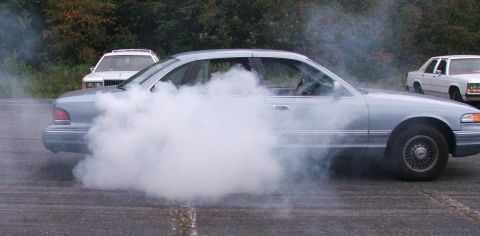 1985 Ford LTD Crown Victoria - Side view of a light blue Ford Crown Victoria sedan mid-burnout, the rear half of the car engulfed in a thick cloud of white tire smoke. Ch…