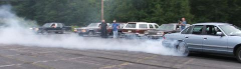 1985 Ford LTD Crown Victoria - A light blue full-size Ford sedan lays a long trail of white tire smoke across a paved lot, the smoke stretching the width of the frame. Sp…