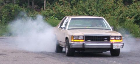 1985 Ford LTD Crown Victoria - Tan 1985 Ford Crown Victoria LTD performing a burnout on a paved surface with trees in the background.
