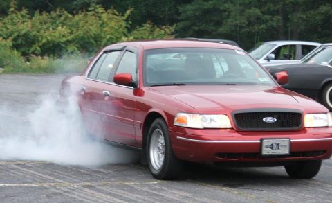 1985 Ford LTD Crown Victoria - A red sedan performs a burnout, creating a cloud of smoke. Other vehicles are visible in the background.