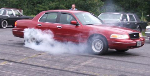 1985 Ford LTD Crown Victoria - A dark red Ford Crown Victoria sedan doing a burnout in a paved lot, tire smoke pouring off the right rear wheel. The car wears chrome wire…