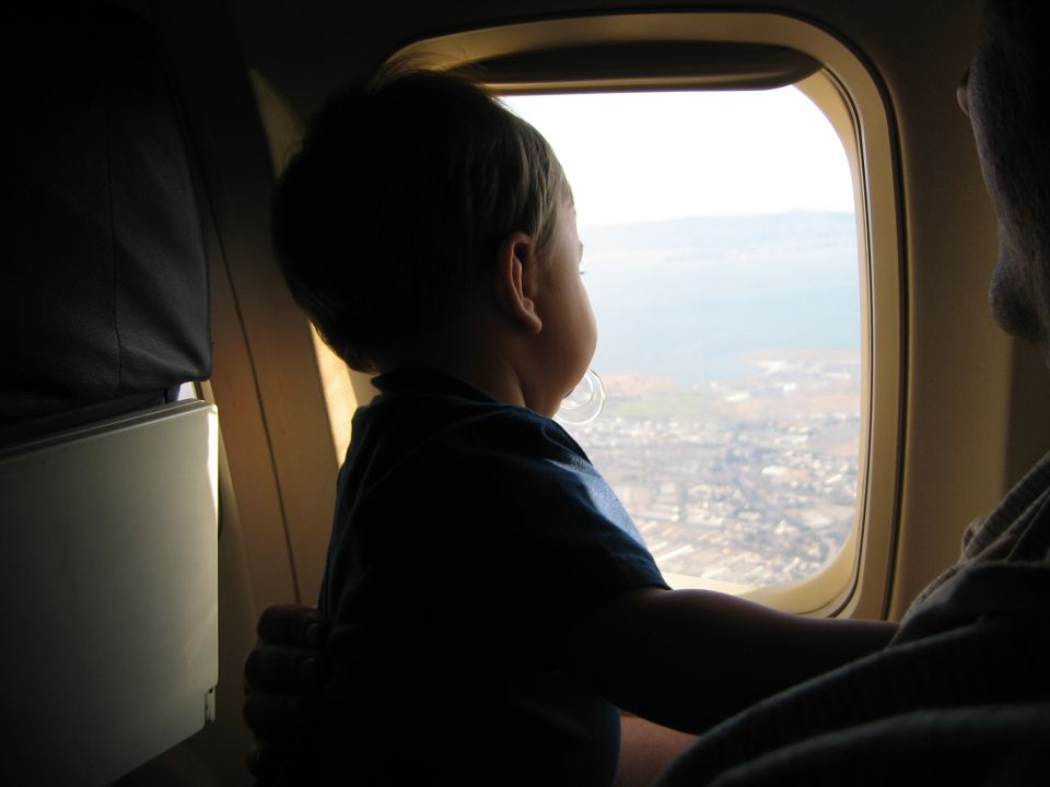Young child looking out an airplane window at the coast below, held by an adult