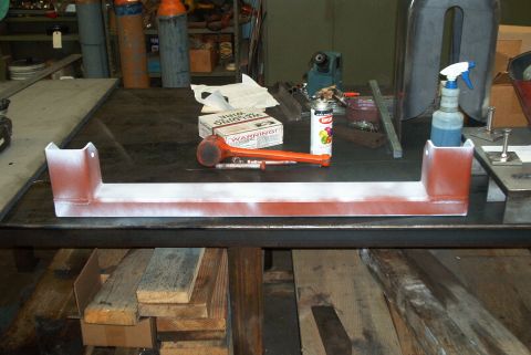 1983 Ford Bronco - A metal crossmember part, painted red and white, sits on a workbench with various tools and supplies nearby.