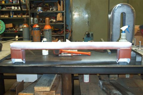 1983 Ford Bronco - A metal crossmember sits on a workbench in a workshop setting, with tools and spray bottles nearby.
