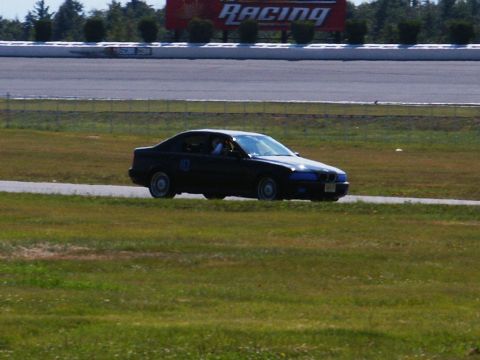 2001 Mercury Grand Marquis - A dark sedan with a racing number on its door drives on a track, with a large 'RACING' sign in the background.
