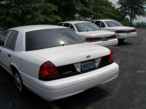 2001 Ford Crown Victoria - Three white Ford Crown Victorias are parked in a lot, showing their rear ends.