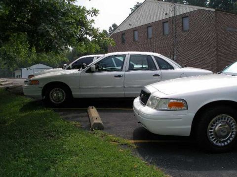 2001 Ford Crown Victoria - Two white Ford Crown Victorias are parked in a lot next to a brick building.