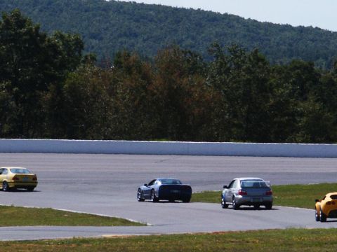 2001 Mercury Grand Marquis - Multiple cars race on a track with a wooded hillside under a clear sky.