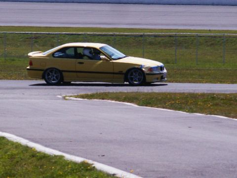 2001 Mercury Grand Marquis - A yellow BMW coupe is captured on a racetrack, with a driver visible inside wearing a helmet.