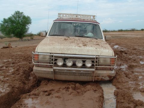 1990 Ford Bronco - A white SUV is stuck in thick mud, its front grille and headlights visible.