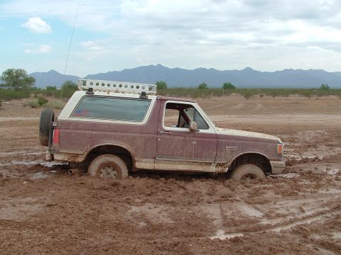 1990 Ford Bronco - Passenger-side profile of a maroon and white two-tone Ford Bronco buried to the axles in thick brown mud on an open desert flat, with mount…