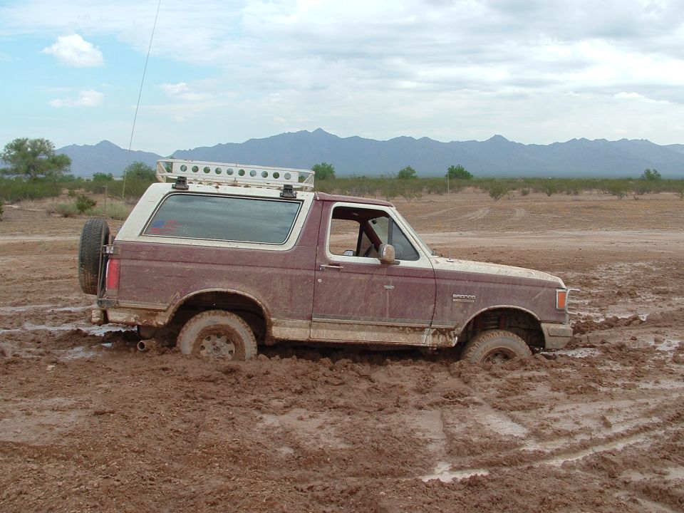 Maroon and white Ford Bronco stuck axle-deep in desert mud with roof light bar and rear tire carrier