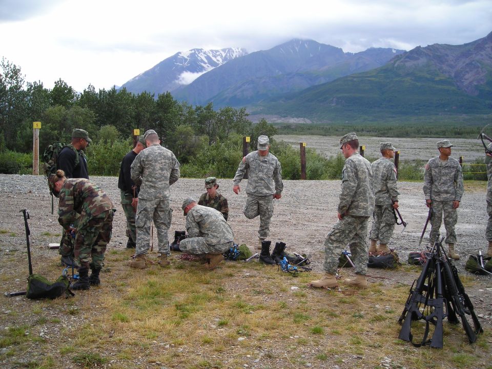 soldiers in camouflage uniforms gather gear and rifles outdoors with mountains in background