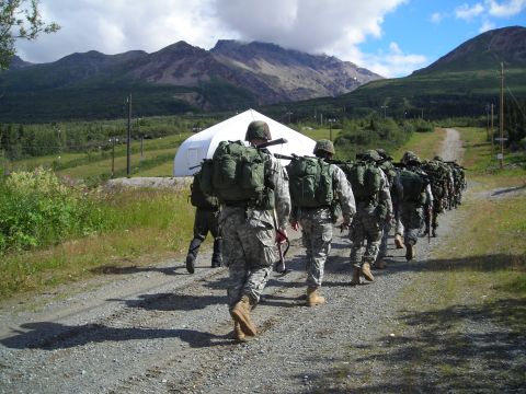 2003 Jeep TJ - A column of soldiers marches down a gravel road towards a large white tent, with a mountainous landscape behind them.