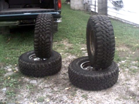 1996 Ford Bronco - Two pairs of off-road tires are stacked on gravel, with the rear of a dark green vehicle visible in the background.