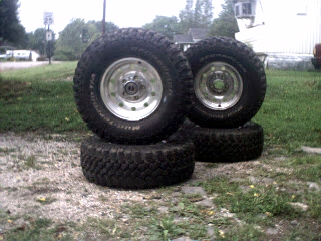 Four off-road tires with chrome rims stacked on grass