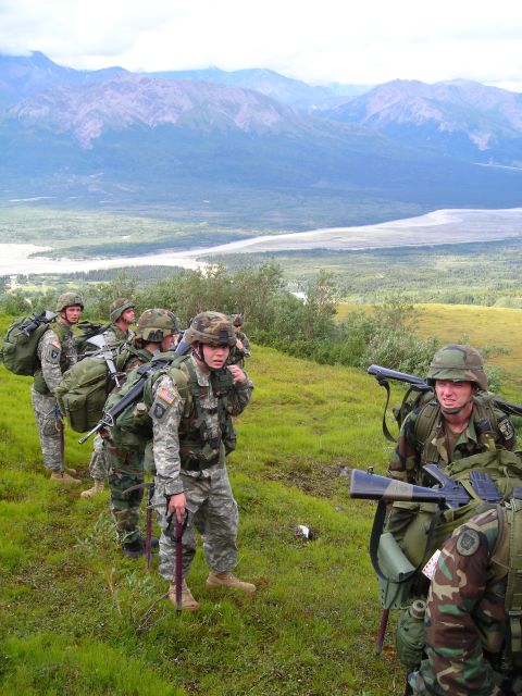 2003 Jeep TJ - A group of soldiers equipped with gear and weapons traverse a grassy hillside under a cloudy sky, with a vast valley and mountains visible…