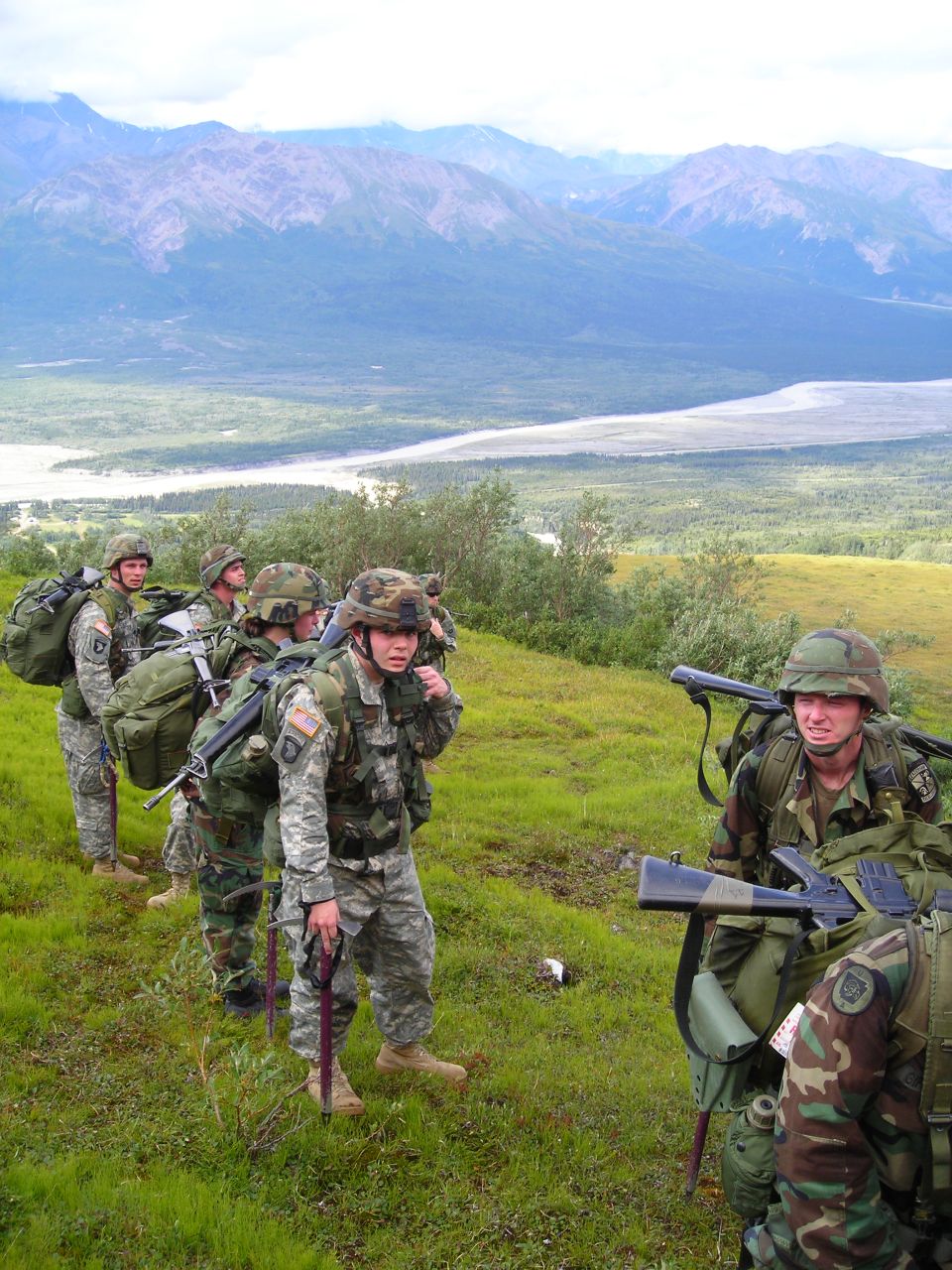 Soldiers in camouflage uniforms with large backpacks and rifles walk single file on a grassy slope with mountains in the background