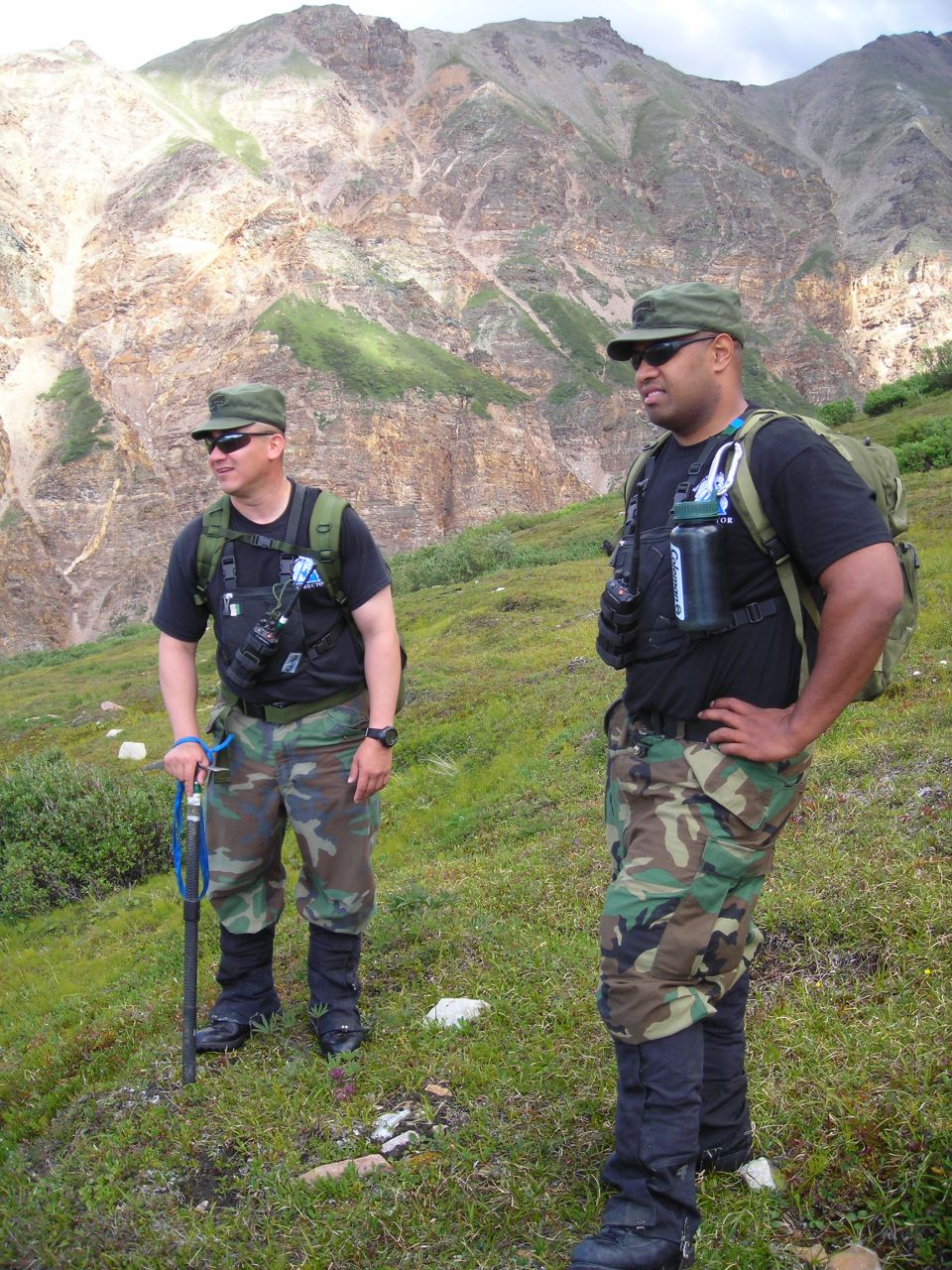 Two men in camouflage stand in a grassy mountain landscape