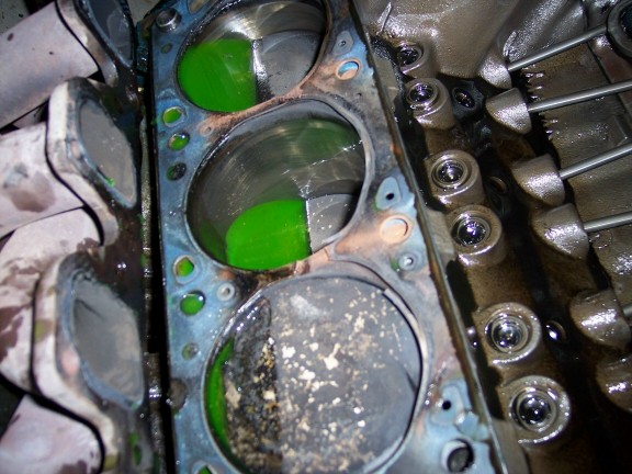 V8 block deck with head removed, green coolant pooled on pistons and old head gasket in place