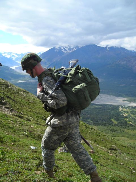 2003 Jeep TJ - A soldier ascends a steep, grassy incline, burdened by a substantial backpack, with a vast valley and mountains stretching out behind.