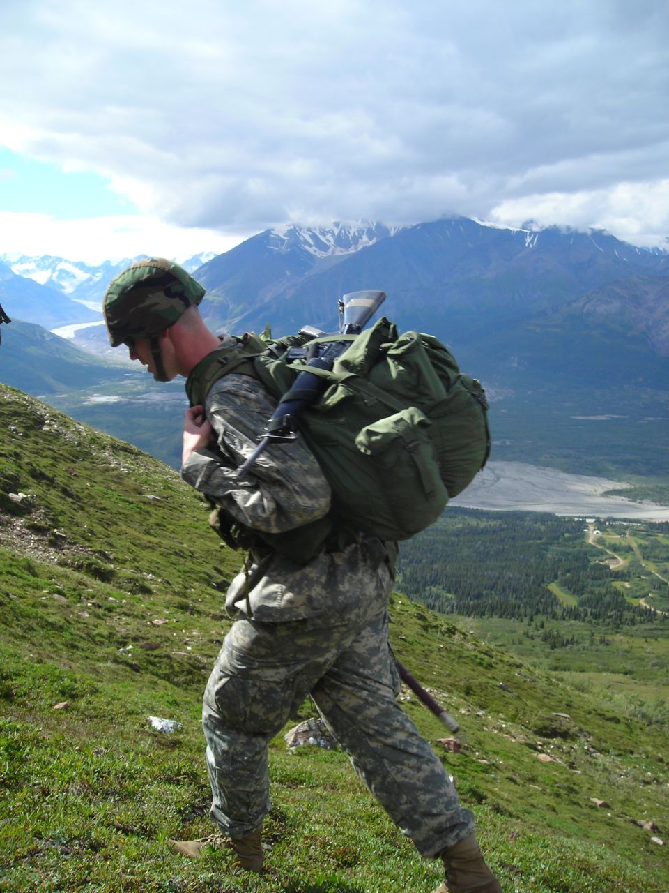 Soldier in camouflage uniform and helmet carrying a large green backpack hikes up a grassy mountain slope with snow-capped mountains in the background
