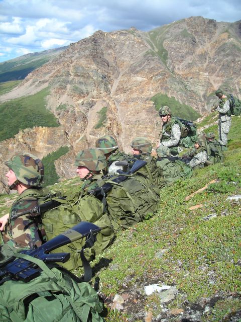 2003 Jeep TJ - Soldiers rest on a hillside with backpacks and rifles, overlooking a mountain landscape under a cloudy sky.
