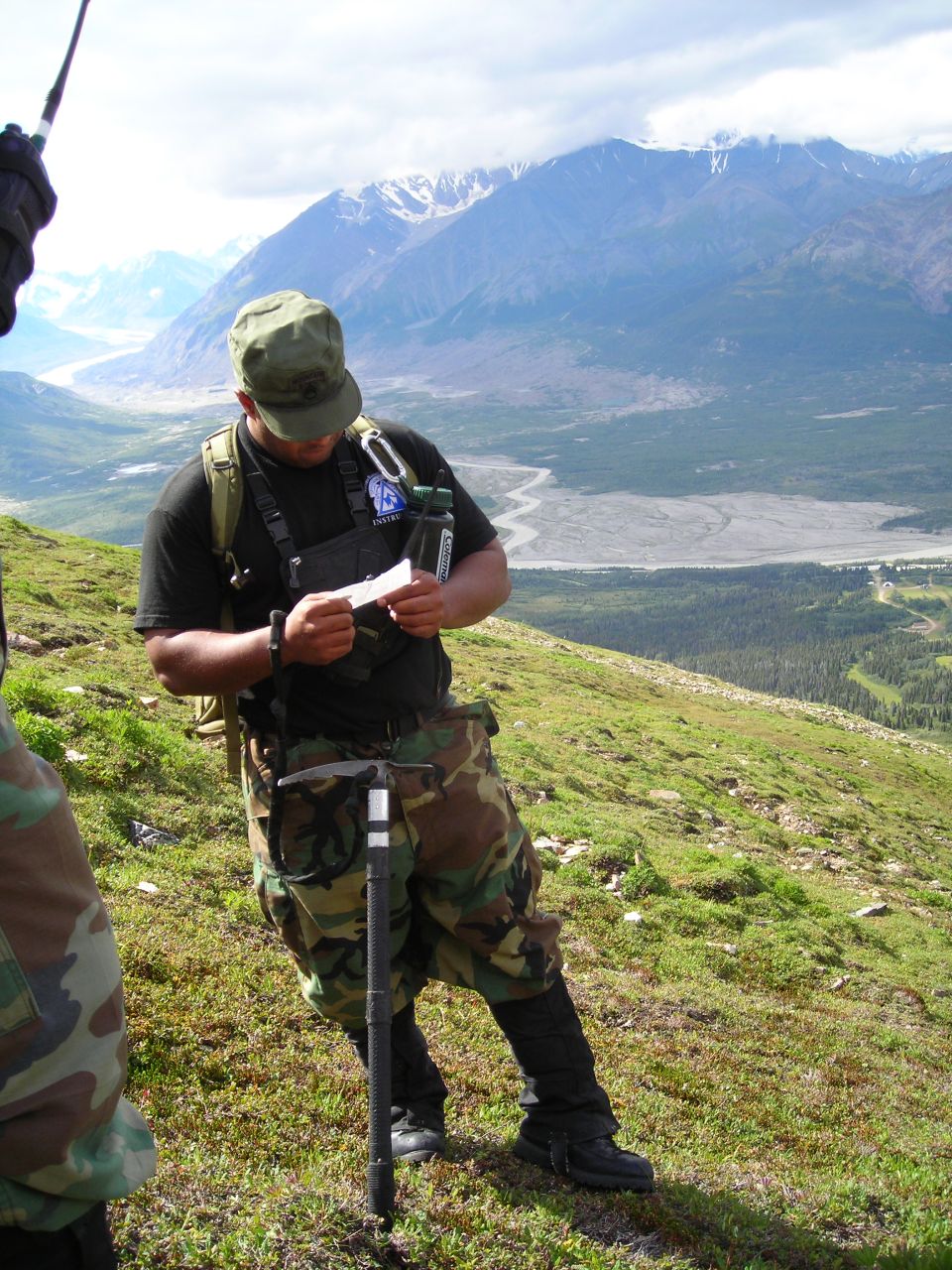 Man in camouflage pants and cap reads paper on grassy mountain slope with snowy mountains in background