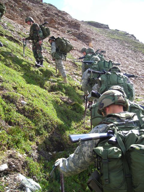 2003 Jeep TJ - A group of soldiers in camouflage gear ascend a steep, vegetated mountainside, carrying large backpacks and rifles.