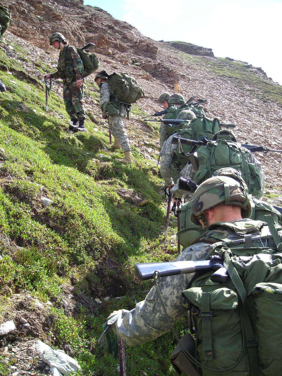 Soldiers hiking up a rocky, grassy slope with backpacks and rifles