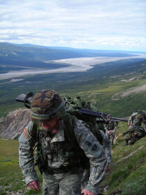 2003 Jeep TJ - Soldiers in camouflage uniforms hike up a grassy mountain slope, carrying rifles and large backpacks, with a wide valley and river visible…