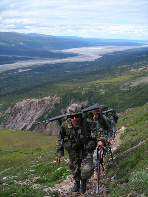 2003 Jeep TJ - Soldiers ascend a mountain trail, carrying rifles and backpacks, with a vast valley and river visible in the distance.