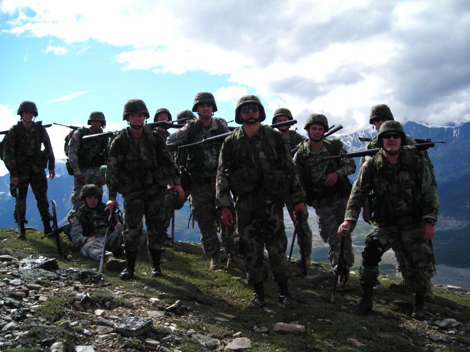 group of soldiers in camouflage uniforms and helmets, carrying rifles, standing on a rocky, grassy hillside with mountains in the background