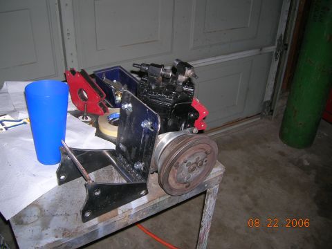 1985 Ford Bronco - An engine compressor part with a pulley is shown on a workbench next to a blue cup and a green gas cylinder.