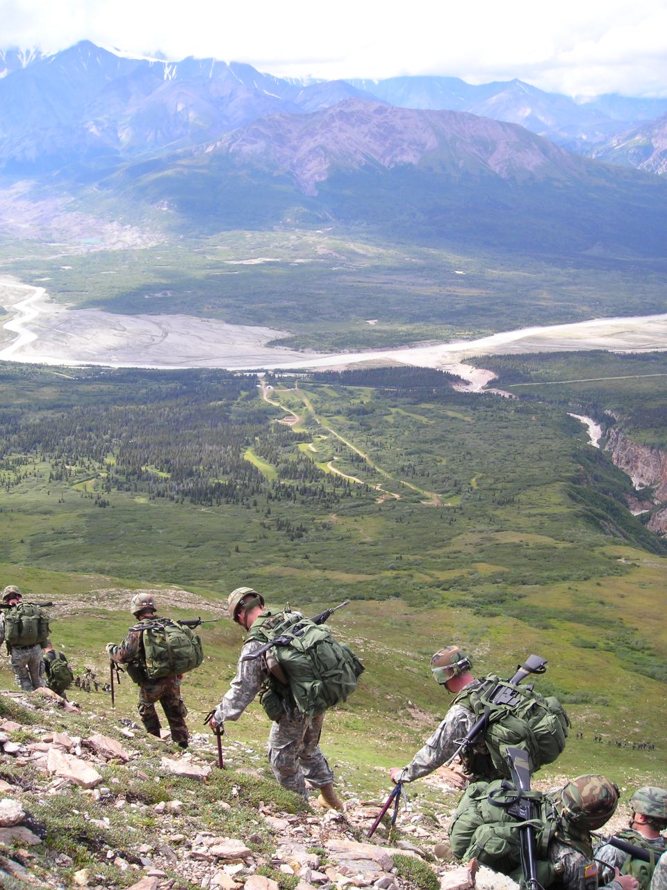 Soldiers with large backpacks hiking up a rocky mountain slope with a valley and mountains in the background