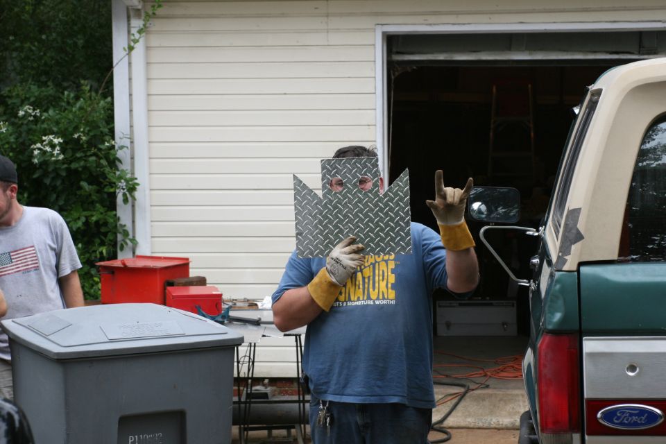 Man holding a diamond-plate shield over his face throwing horns outside a garage