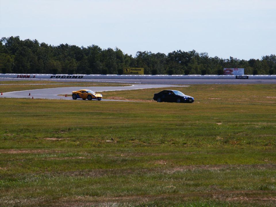 Yellow sports car and dark sedan on a racetrack with trees and sky in the background