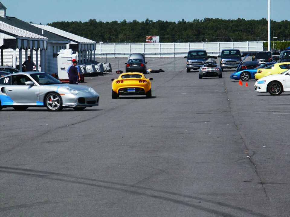 Silver Porsche 911 with blue accents and number 18, yellow Lotus Elise, and other cars on a paved lot