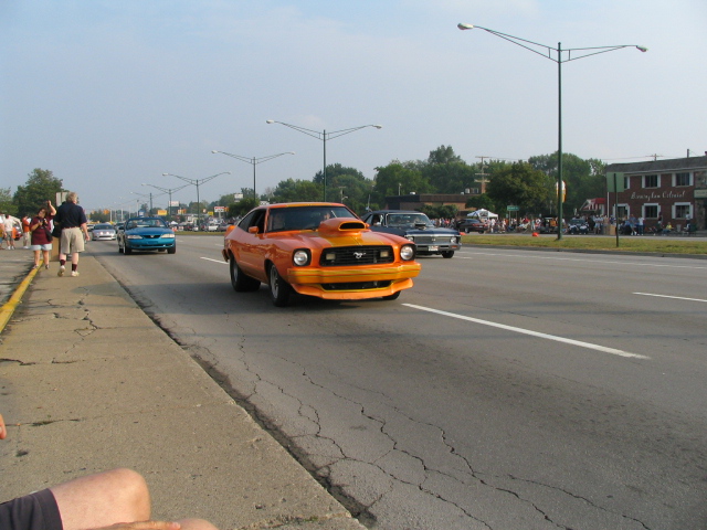 Orange Mustang II Cobra cruising a multi-lane road during a street cruise event