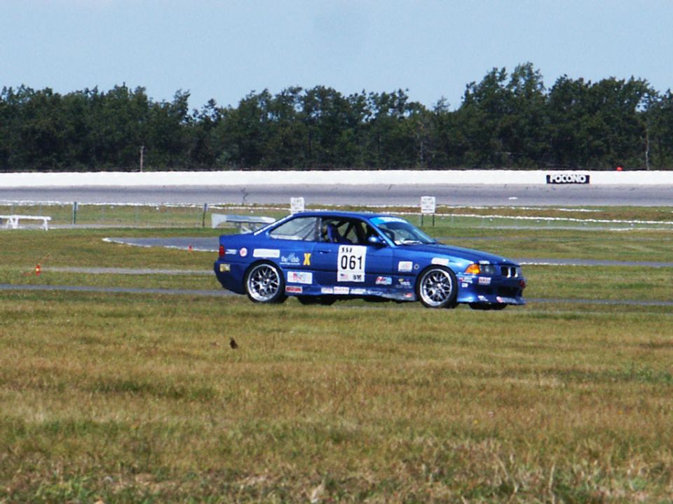 Blue BMW E36 race car with number 061 on door, on a grassy track infield