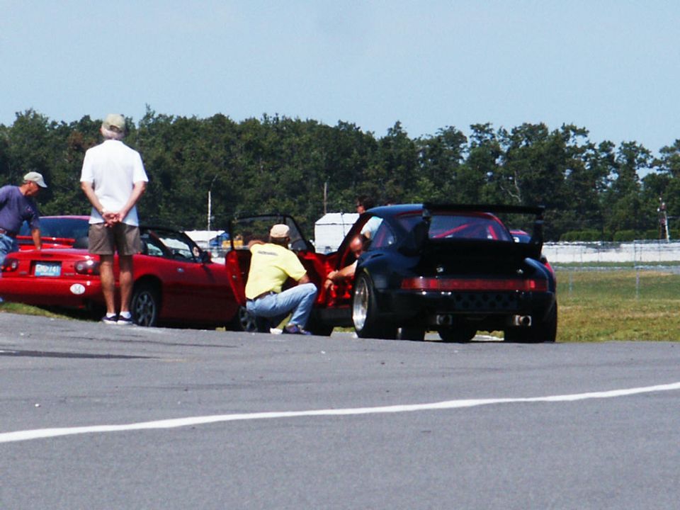 Black Porsche 911 with a large rear spoiler parked next to a red convertible. People are near the cars