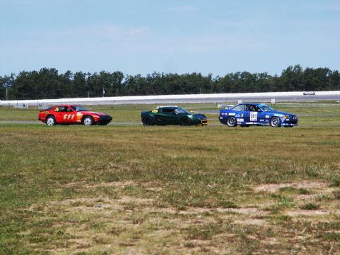 2001 Mercury Grand Marquis - Three cars, a red Porsche, a green Lotus, and a blue BMW, compete on a race track under a clear sky.