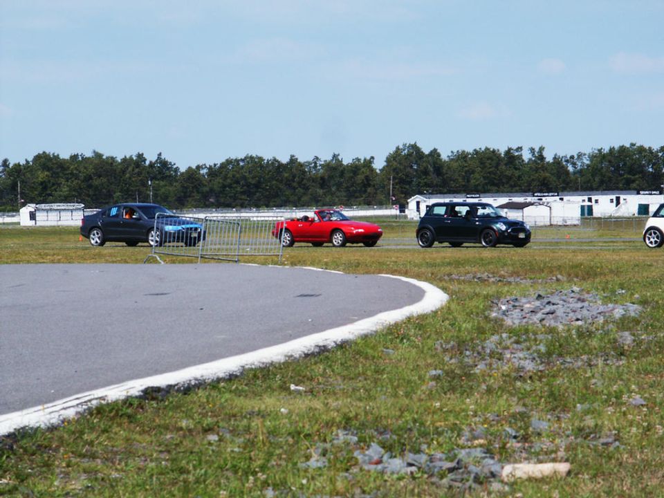 Several cars, including a red convertible and a black mini, on a racetrack with trees and buildings in the background