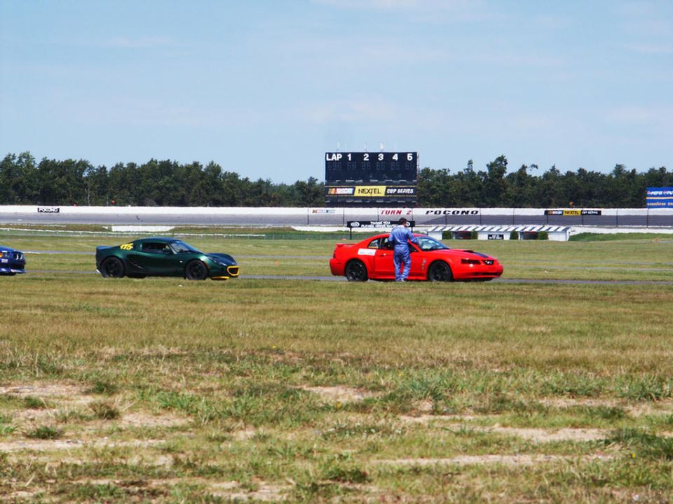 Red sports car on a race track, driver standing by the open door. Green sports car and blue car visible in background